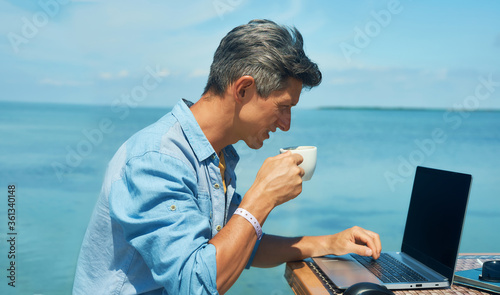man freelancer working at morning on the beach by the sea, drinking coffee, using laptop computer. dream office job workplace. Freelance concept