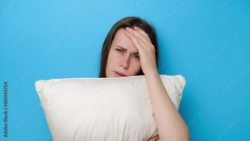 Discontent young woman has gloomy face, carries white pillow, has insomnia, cant get asleep late, isolated on blue background. Lady stressed because of too early wakeup, suffering from lack of sleep Discontent young woman has gloomy face, carries white pillow, has insomnia, cant get asleep late, isolated on blue background. Lady stressed because of too early wakeup, suffering from lack of sleep