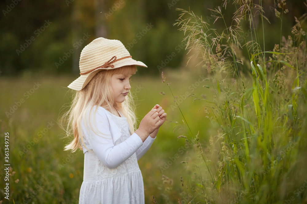 Close-up portrait of  cute little blonde girl in straw hat and white dress outdoors in a park or field next to grass blades. Sunny summer evening. The atmosphere of summer time, childhood, happiness.
