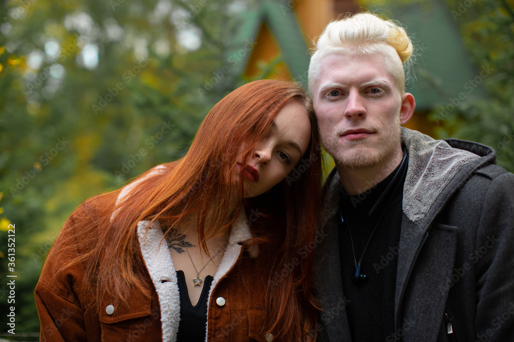 Portrait of an unusual couple. An albino man in a dark jacket and a girl with bright red hair in a corduroy jacket look to the camera against a backdrop of green trees