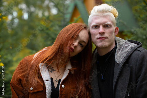 Portrait of an unusual couple. An albino man in a dark jacket and a girl with bright red hair in a corduroy jacket look to the camera against a backdrop of green trees