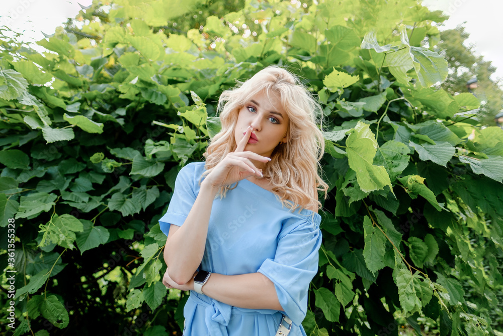 Young beautiful blonde girl in a blue dress bites her finger and touches her lips with her hands. Naughty, funny and playful woman in the park. Young beautiful blonde girl in a blue dress bites her finger and touches her lips with her hands. Naughty, funny and playful woman in the park.