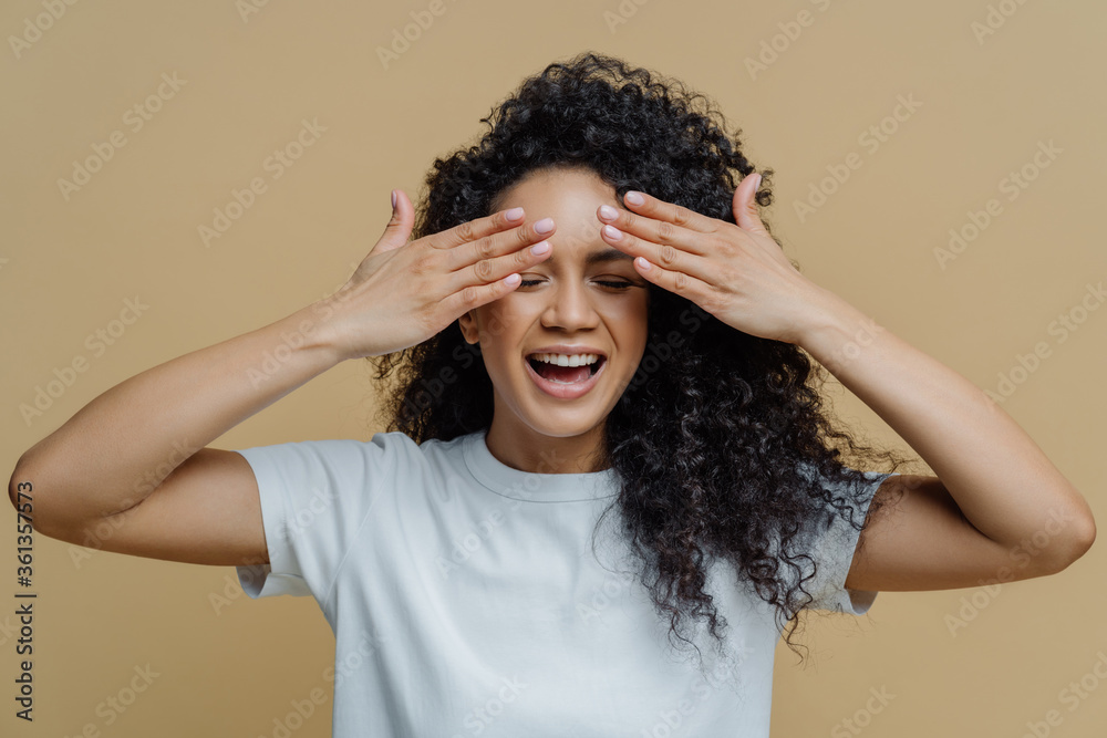 Horizontal shot of happy dark skinned woman covers eyes and smiles, has fun and hides face, has curly hair, dressed in casual white t shirt, isolated on brown background, waits for surprise. Horizontal shot of happy dark skinned woman covers eyes and smiles, has fun and hides face, has curly hair, dressed in casual white t shirt, isolated on brown background, waits for surprise.