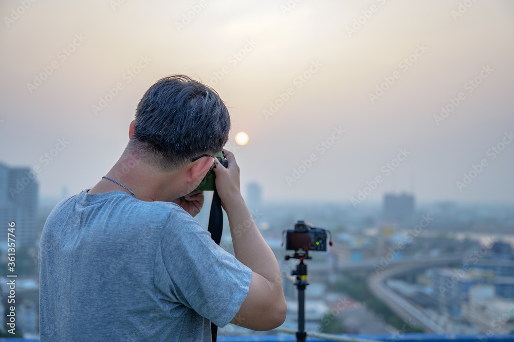 Asian photographer man is using his Dslr or mirrorless camera to take pictures of cityscape with the Sunrise or sunset