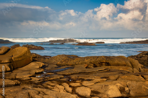 A landscape with rocks in the foreground and waves breaking over rocks in the background. Rain clouds forming on the horizon.