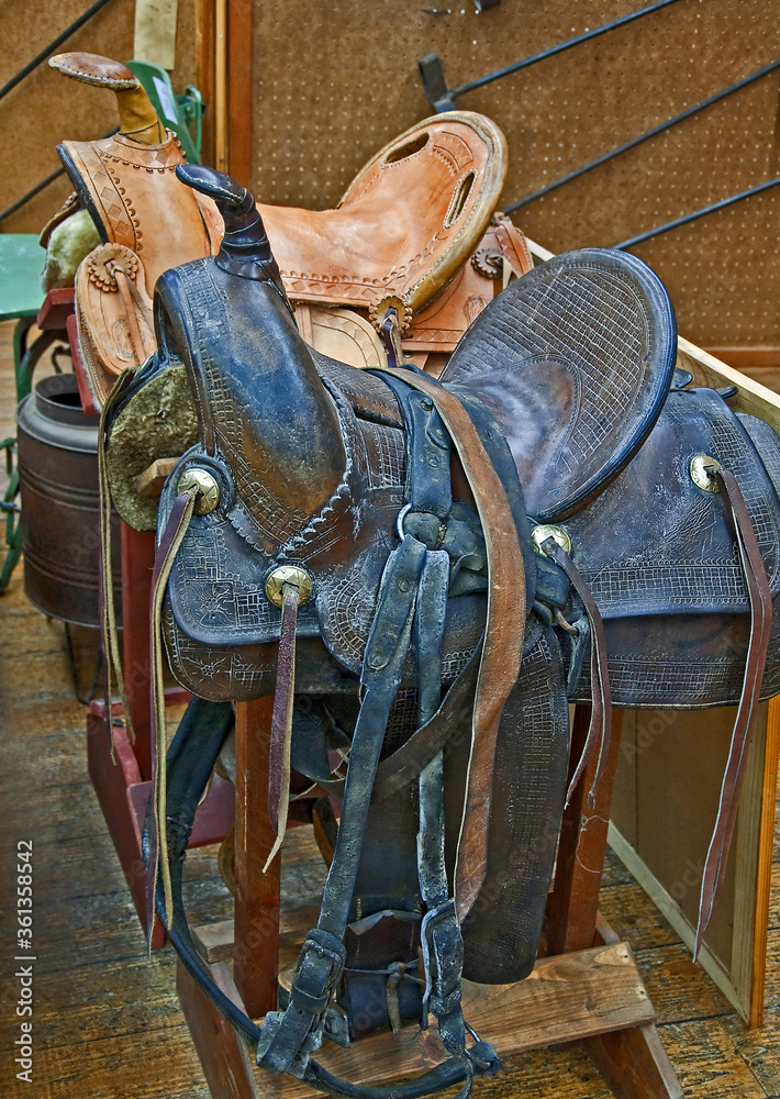 Two retro leather horse saddles, one brown and the other a light tan color, on sawhorses with various iron brands in the background.