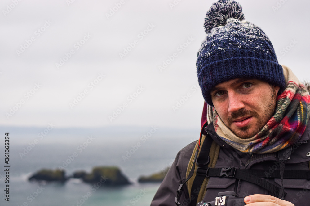 Photo of a young and attractive man with beard standing in the nature with winter clothes and a backpack in the Giants Causeway, Belfast. Holidays concept