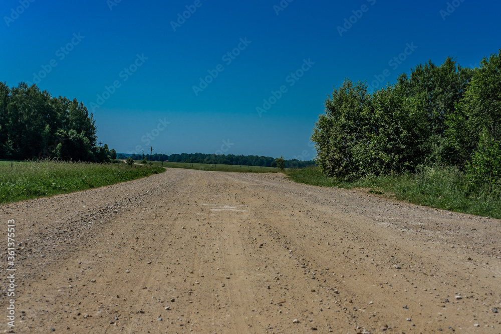 A deserted dirt road made of sand and small stones, extending into the distance, surrounded by grass and shrubs, against a blue sky.