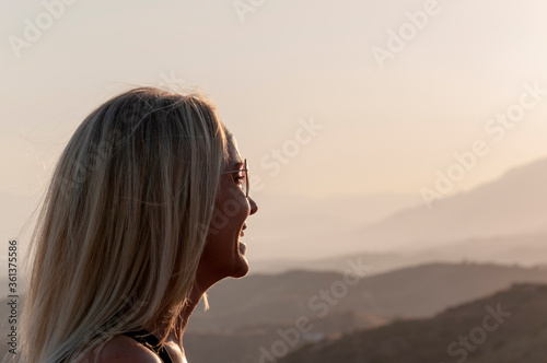 Blonde girl in sunglasses laughs on a blurred background of mountains silhouettes, profile shot