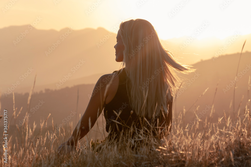 A girl sits in the grass on a hilltop with her eyes closed and meditates against the backdrop of mountain peaks and the sunset, her blond hair flying in the wind.