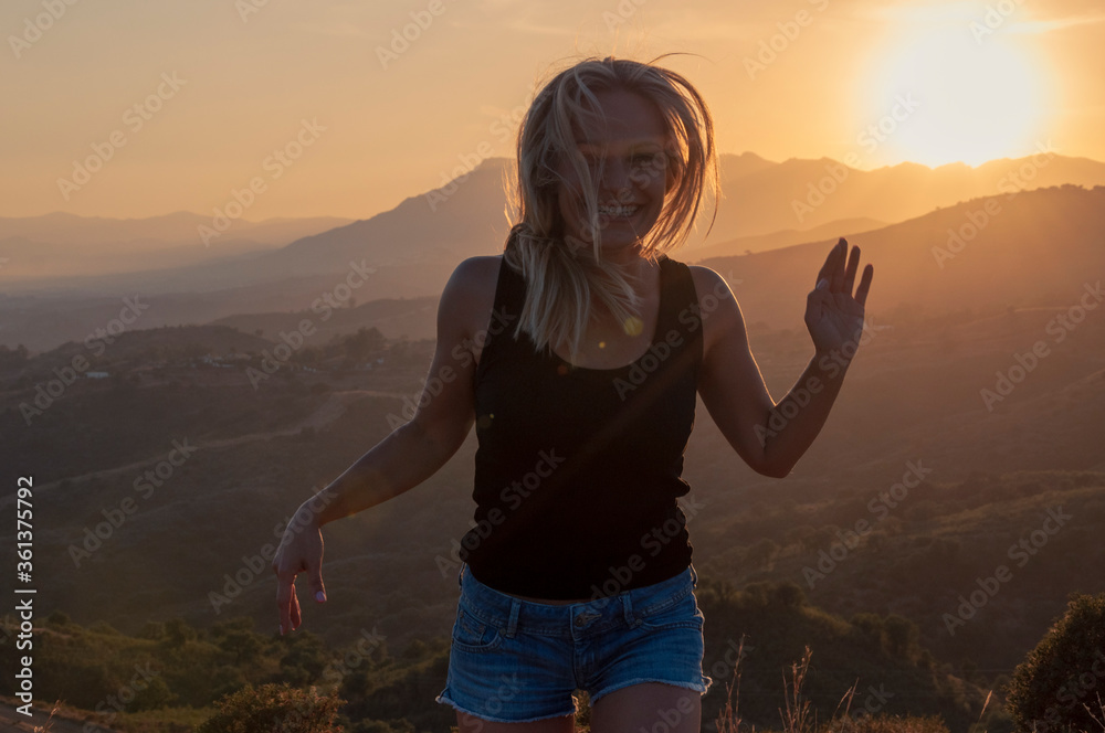 Girl with tousled hair in the wind in sneakers, a T-shirt and shorts laughs and waves her hand against the backdrop of the mountains during sunset