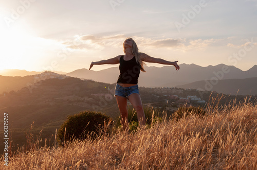 A blonde girl stands with arms spread out to the side in the grass on a hilltop against the backdrop of the mountains during sunset © Andrei