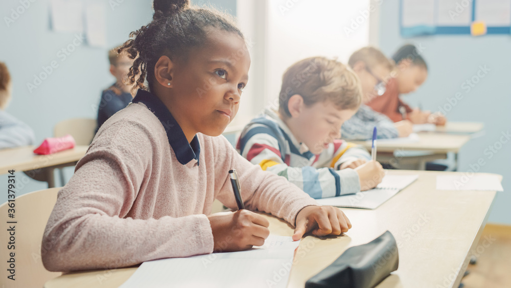 In Elementary School Classroom Brilliant Black Girl Writes in Exercise Notebook, Taking Test and Writing Exam. Junior Classroom with Diverse Group of Children Working Diligently and Learning New Stuff In Elementary School Classroom Brilliant Black Girl Writes in Exercise Notebook, Taking Test and Writing Exam. Junior Classroom with Diverse Group of Children Working Diligently and Learning New Stuff