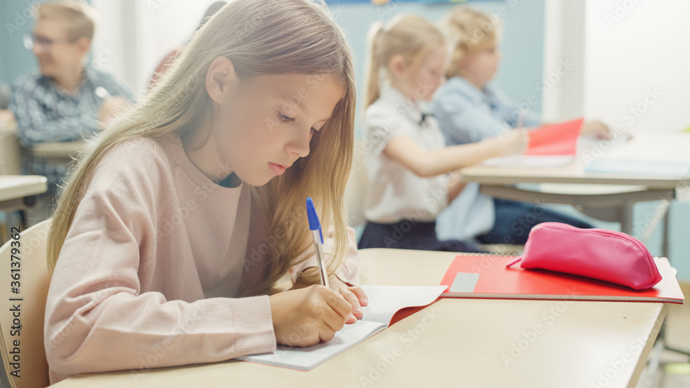 In Elementary School Classroom Caucasian Girl Writes in Exercise Notebook, Taking Test and Writing Exam. Junior Classroom with Diverse Group of Children Working Diligently and Learning New Stuff In Elementary School Classroom Caucasian Girl Writes in Exercise Notebook, Taking Test and Writing Exam. Junior Classroom with Diverse Group of Children Working Diligently and Learning New Stuff
