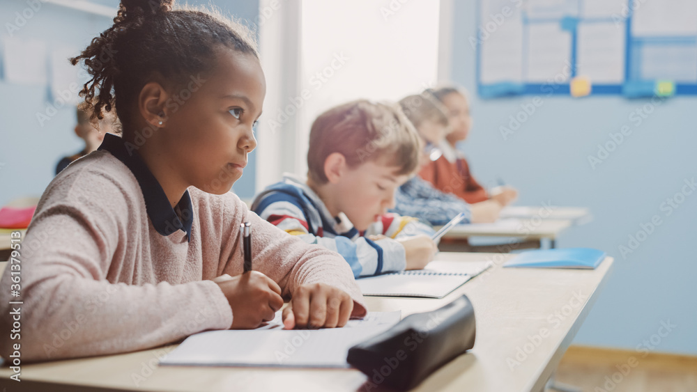 In Elementary School Classroom Brilliant Black Girl Writes in Exercise Notebook, Taking Test. Junior Classroom with Diverse Group of Bright Children Working Diligently and Learning. Side View Portrait In Elementary School Classroom Brilliant Black Girl Writes in Exercise Notebook, Taking Test. Junior Classroom with Diverse Group of Bright Children Working Diligently and Learning. Side View Portrait