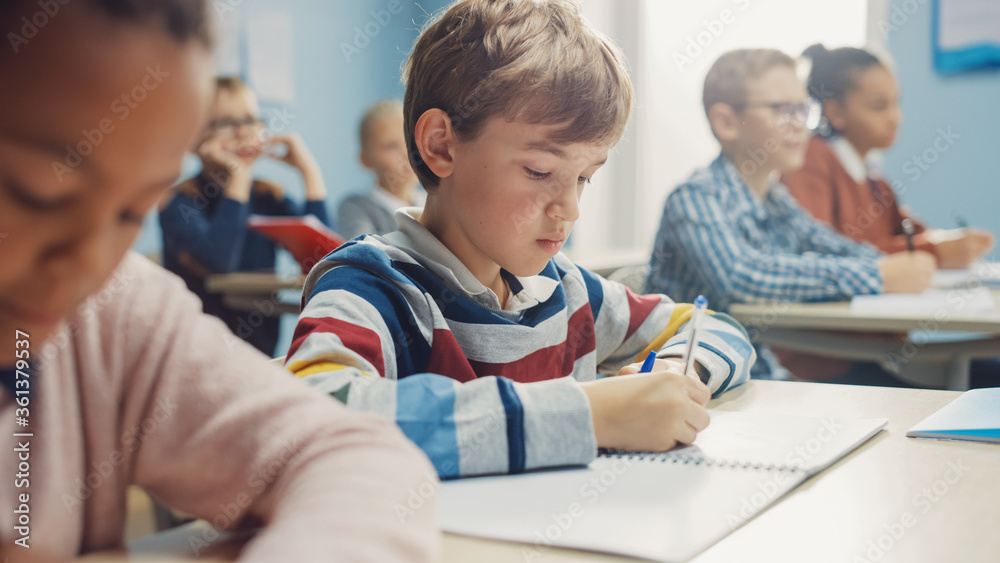 In Elementary School Class: Portrait of Brilliant Caucasian Boy Writes in Exercise Notebook, Taking Test and Writing Exam. Diverse Group of Bright Children Working Diligently and Learning In Elementary School Class: Portrait of Brilliant Caucasian Boy Writes in Exercise Notebook, Taking Test and Writing Exam. Diverse Group of Bright Children Working Diligently and Learning