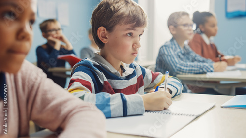 In Elementary School Class: Portrait of Brilliant Caucasian Boy Writes in Exercise Notebook, Taking Test and Writing Exam. Diverse Group of Bright Children Working Diligently and Learning photo