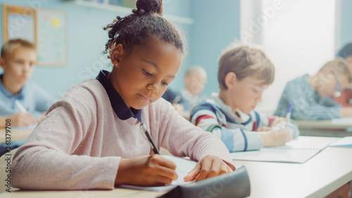In Elementary School Class: Portrait of a Brilliant Black Girl with Braces Writes in Exercise Notebook, Smiles. Junior Classroom with Diverse Group of Children Learning New Stuff