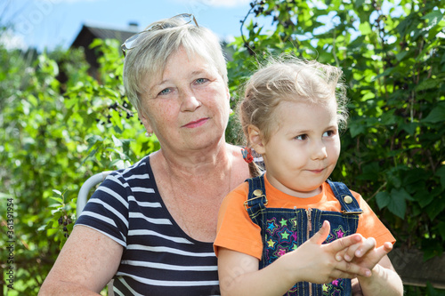 Portrait of little granddaughter and her grandmother in summer garden between green plants