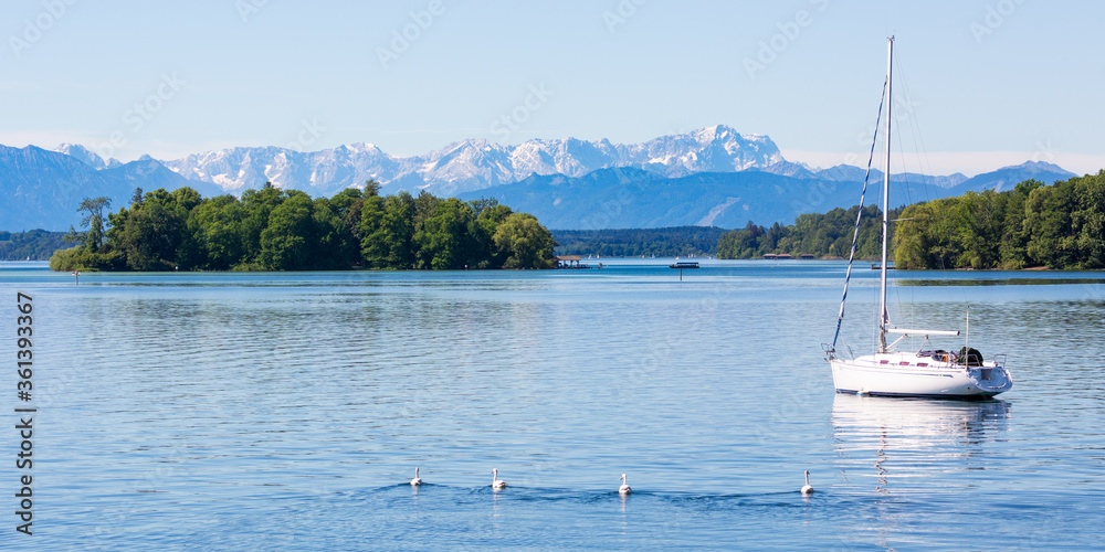 Scenic landscape of Lake Starnberg with Roseninsel (rose island) and an anchoring sailboat. Snow capped mountains at the horizon.