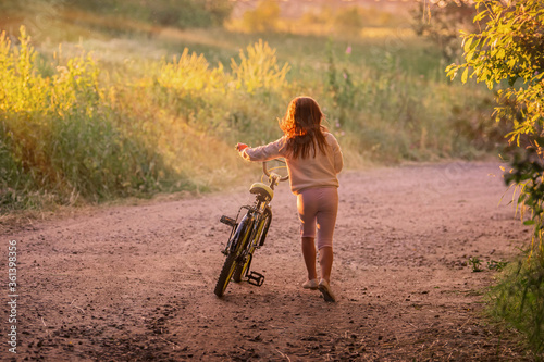 little girl goes with a bicycle on a rural road in nature at sunset