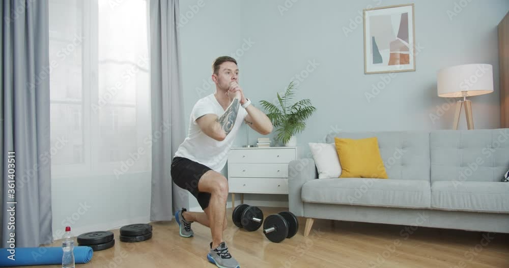 Young fitness trainer stretching, warming up, working out at home. A sporty muscular guy performing lower body exercise, jumping lunges in the living room. Self-motivation, healthcare, sport concept. Young fitness trainer stretching, warming up, working out at home. A sporty muscular guy performing lower body exercise, jumping lunges in the living room. Self-motivation, healthcare, sport concept.