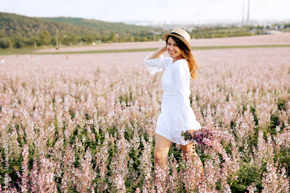 Young brunette woman wearing a straw hat and a white dress, running along a field with pink sage flowers at sunset. Beautiful girl relaxing outdoors, having fun, holding plant. Young brunette woman wearing a straw hat and a white dress, running along a field with pink sage flowers at sunset. Beautiful girl relaxing outdoors, having fun, holding plant.