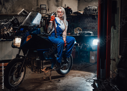 Tattooed hipster girl in work overalls hold big wrench while sitting on sportbike in garage or workshop, smiling and looking on camera