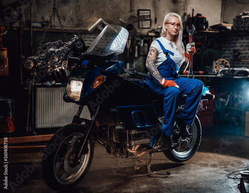 Young blond woman mechanic in work overalls hold big wrench while sitting on sportbike in garage or workshop