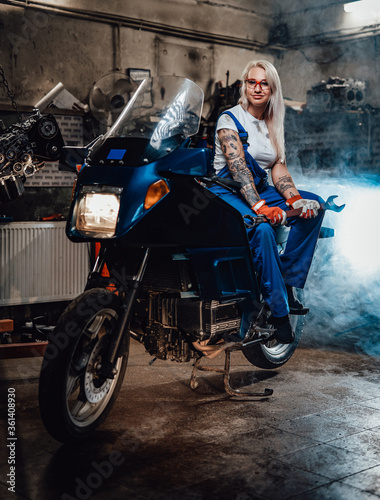 Young blond woman mechanic in work overalls hold big wrench while sitting on sportbike in garage or workshop, smiling and looking on camera