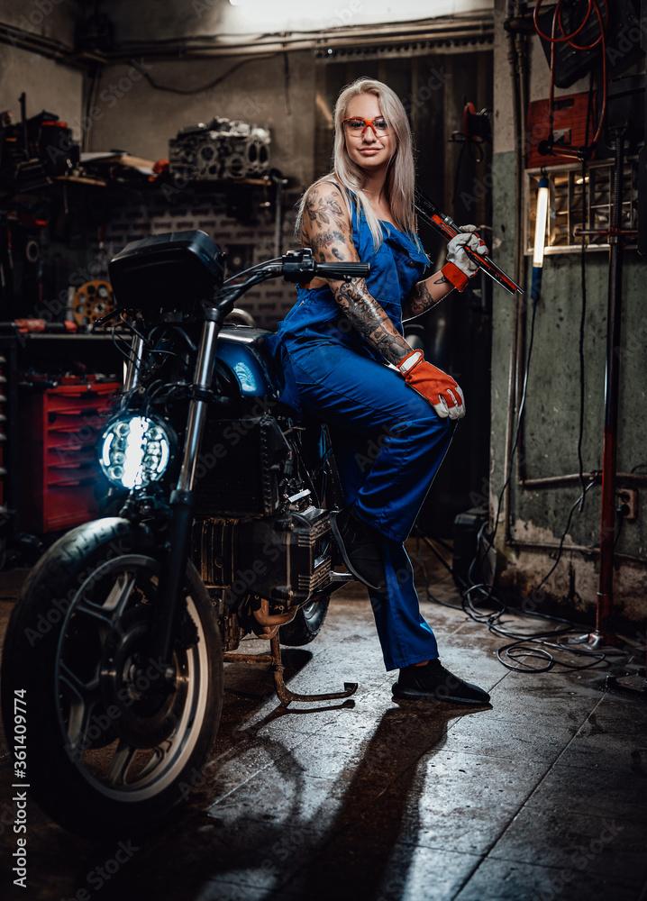Tattooed hipster girl in work overalls hold a big wrench while leaning on her naked bike in garage or workshop, smiling and looking on camera