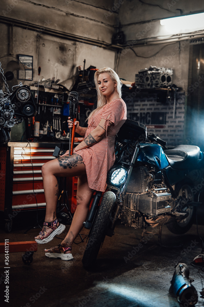 Beautiful blonde girl with tattooed body wearing pink dress posing next to the suspended flat engine and naked bike in garage or workshop
