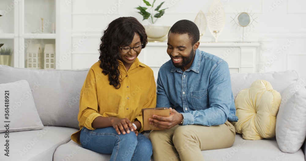 Happy African American couple husband and wife sitting on sofa using tablet at home.