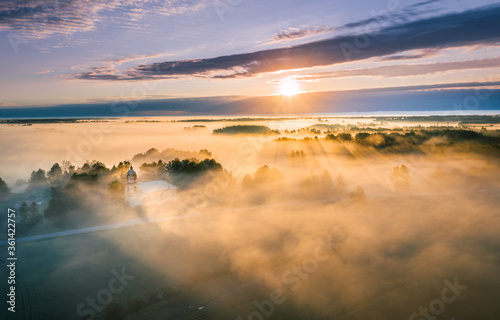 Majestic church covered in fog and surrounded by forest. Aerial view over picturesque countryside with at sunrise with long light rays. 