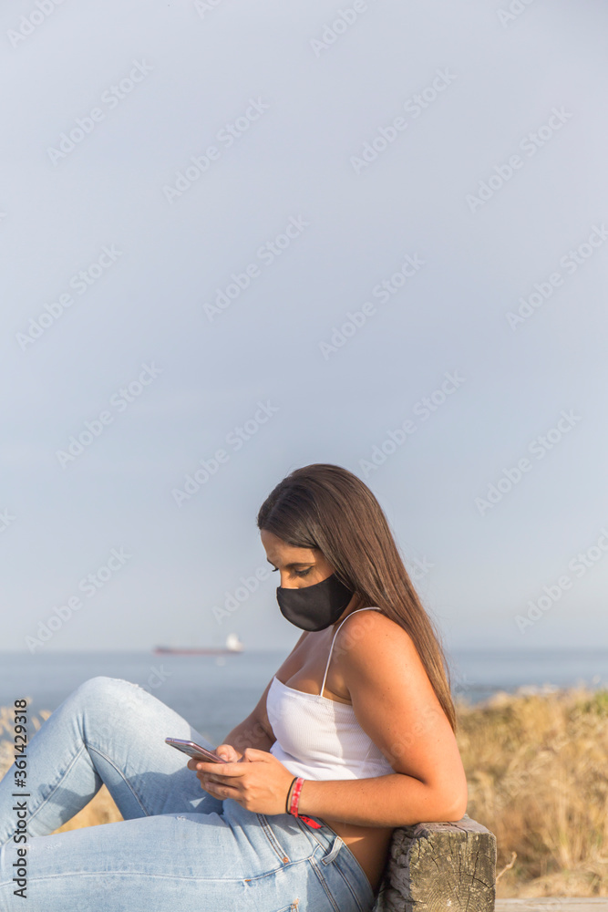 young woman with mask looks at her phone sitting on a bench overlooking the sea