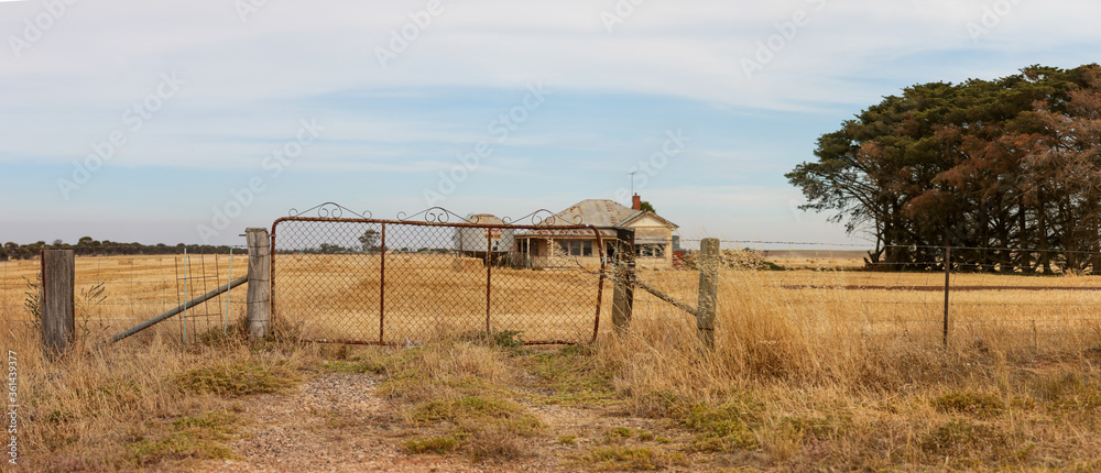 Fototapeta premium Panoramic image of an old farm gate and a timber worn out abandoned traditional Australian farm house in the middle of a newly harvested field on a agricultural property in rural Victoria, Australia