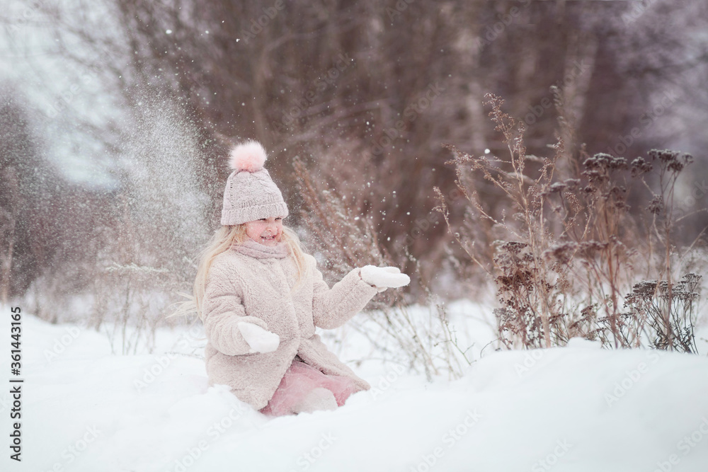 Cute little girl child blonde in a pink knitted hat with a pompom and a fur coat sitting in a snowdrift in the winter outdoors throwing snow. Winter walks, fun outdoors winter time.