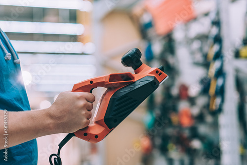 A man holds in his hand an electric planer for a tree. A buyer in a hardware store selects a product.