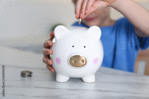 Little boy putting coin into piggy bank at marble table indoors, closeup