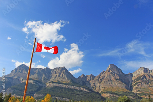Canadian national flag with maple leaf symbol hanging in the wind. Rocky mountains and blue sky in the background.