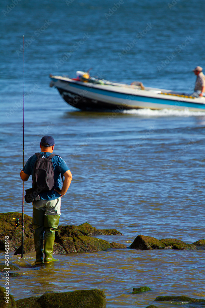 A fisherman wearing long fishing boots , a backpack and a hat is standing on the rock with a long fishing rod. Another fisherman is passing by on a small motor boat in the background