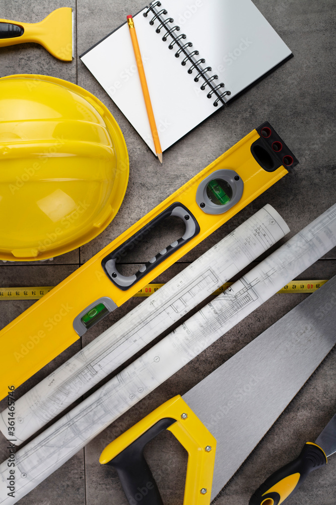 Contractor concept. Tool kit of the contractor: yellow hardhat, libella, hand saw. Plans and notebook on the gray tiles background. Contractor concept. Tool kit of the contractor: yellow hardhat, libella, hand saw. Plans and notebook on the gray tiles background.
