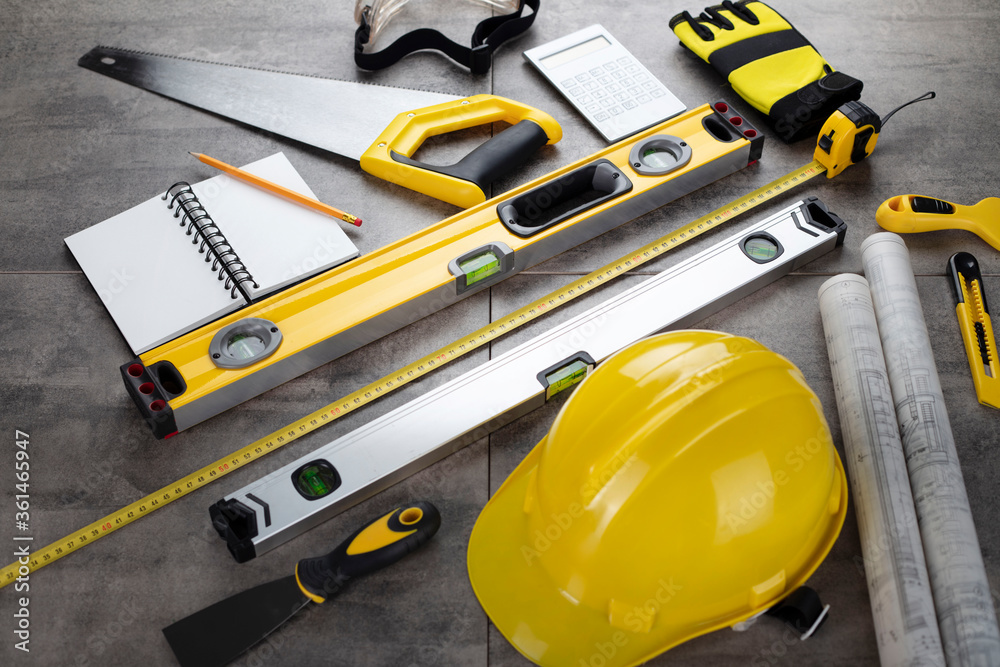 Contractor concept. Tool kit of the contractor: yellow hardhat, libella, hand saw. Plans and notebook on the gray tiles background.