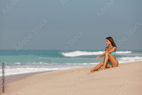 Young beautiful girl sits on the sandy beach with torquise water and blue sky  during vacation  a trip to a tropical resort. Seascape view  summer vacation concept