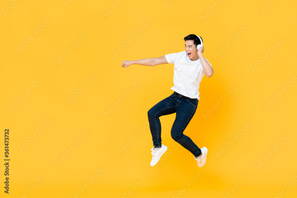 Happy handsome young Asian man wearing headphones punching and jumping in colorful yellow background Happy handsome young Asian man wearing headphones punching and jumping in colorful yellow background