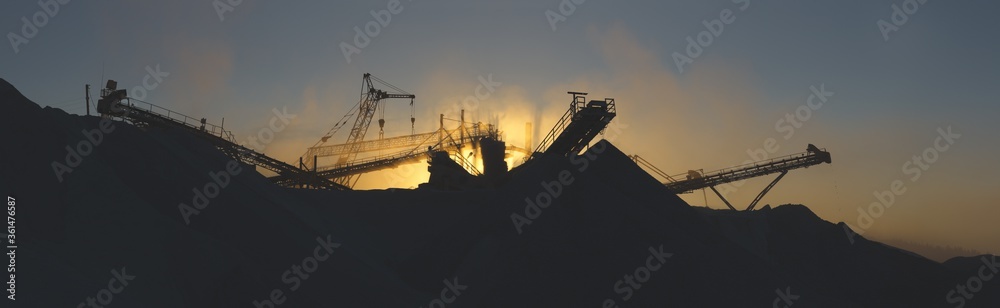 Panorama of the crushing plant equipment, silhouette in a cloud of dust highlighted by evening lighting in orange tones. Mining industry.