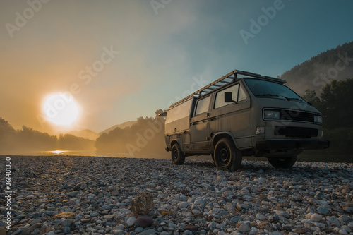 Vintage retro campervan parked on the wild camping ground next to a river at early hazy morning hours. Adventure time for campers in the morning.