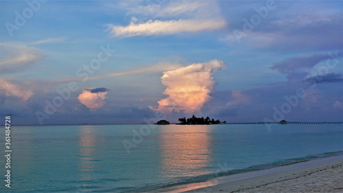 Magical sunset in the Maldives. The aquamarine ocean is calm. The colors of the sky are azure, blue, lilac. Clouds glow in the rays of the setting sun in gold and pink. Reflection in water. 