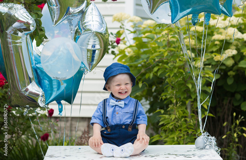 A beautiful little boy kid gentleman in a retro suit with suspenders and a bow tie is sitting on a white tablen. Children's party with balloons happy birthday, 1 year. photo