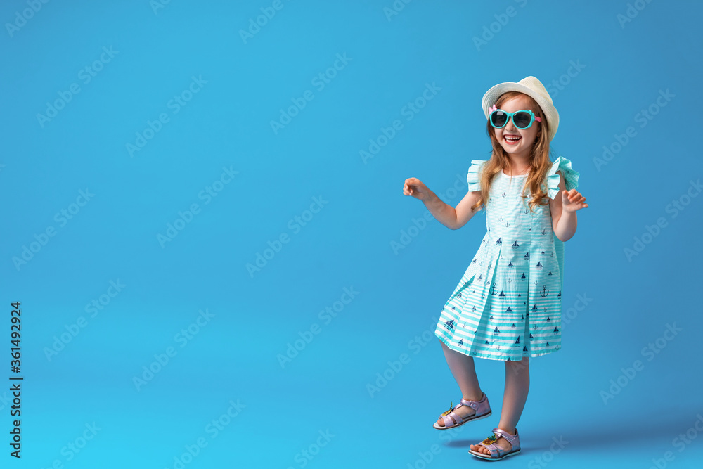 Fototapeta premium cute little girl in a dress, hat and sunglasses poses on a blue background